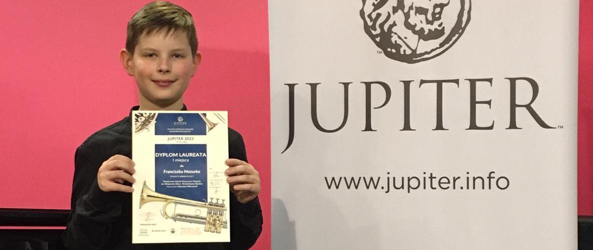 Zdjęcie przedstawia chłopca trzymającego dyplom z konkursu, po prawej duży baner z napisem jupiter Music Info