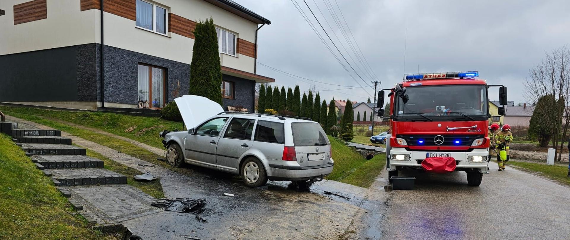 Na zdjęciu pokazano samochód po uderzeniu w przepust drogowy stojący z otwartą klapą silnika na podjeździe do budynku. Na ulicy stoi samochód pożarniczy, a przy nim strażacy.