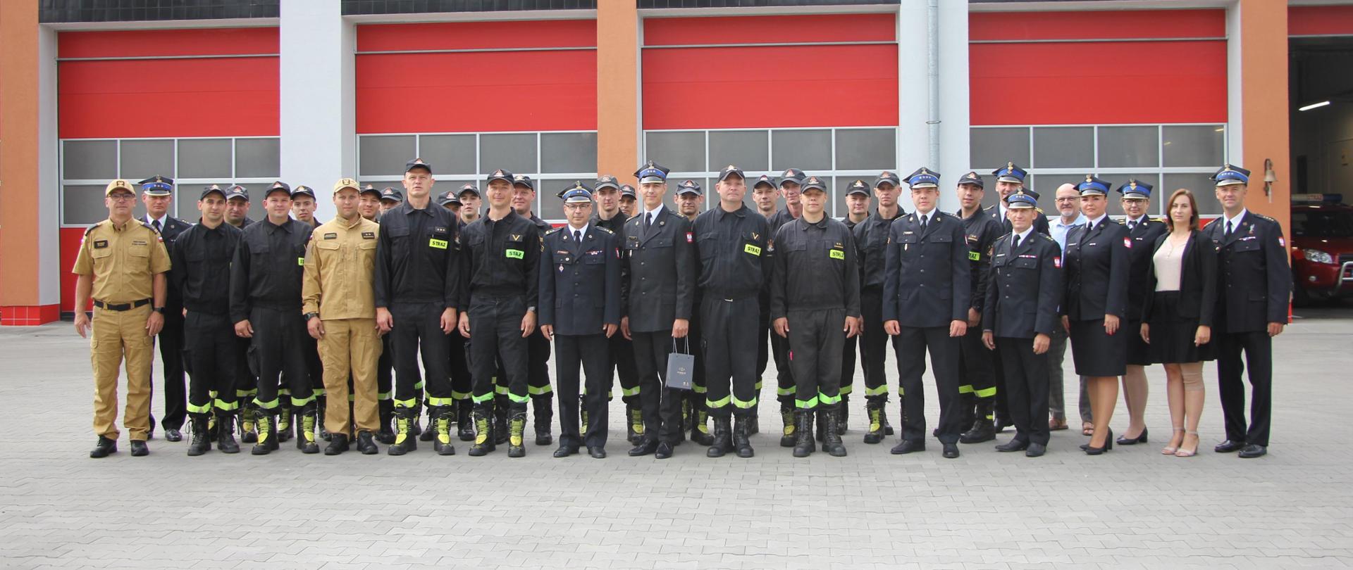 Strażacy i osoby cywilne stoją w dwuszeregu przed budynkiem komendy.