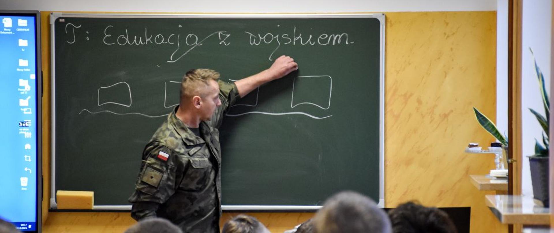 Żołnierz zapisuje na tablicy szkolnej temat lekcji: Edukacja z wojskiem