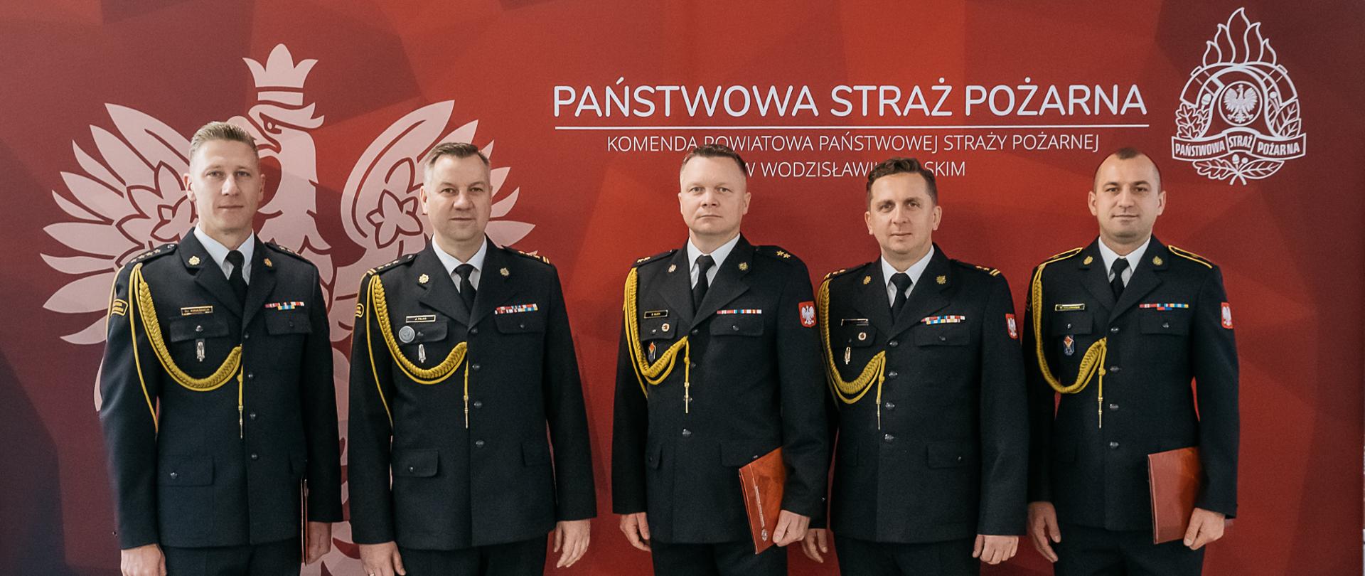 Wspólne zdjęcie komendanta wraz z powołanymi i mianowanymi tego dnia funkcjonariuszami, w tle logo PSP