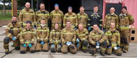 W dwóch rzędach ustawieni są strażacy w piaskowych ubraniach specjalnych, pierwszy rząd klęczy drugi stoi. W tle kontener metalowy