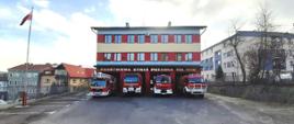 Fotografia kolorowa przedstawia widok trzykondygnacyjnego budynku Komendy Powiatowej Państwowej Straży Pożarnej w Brzozowie. Na pierwszym planie asfaltowy plac manewrowy przed budynkiem. Po lewej stronie maszt flagowy z biało-czerwona flagą na szczycie. Przed budynkiem stoją częściowo w bramach garażowych cztery czerwone pożarnicze pojazdy ciężarowe - od lewej - samochód specjalny podnośnik hydrauliczny, następnie trzy pojazdy ratowniczo-gaśnicze. Wszystkie pojazdy mają włączone białe światła mijania i niebieskie światła błyskowe. Drugi plan wypełnia ściana frontowa budynku Komendy Powiatowej Państwowej Straży Pożarnej w Brzozowie, malowana pasami koloru czerwonego, niebieskiego i żółtego, w okresie jesiennym. Na ścianie parteru napis o treści Państwowa Straż Pożarna Tel. 998. Na parterze widoczne cztery bramy wyjazdowe, w których stoją opisane wcześniej samochody, na pozostałych piętrach okna pomieszczeń.