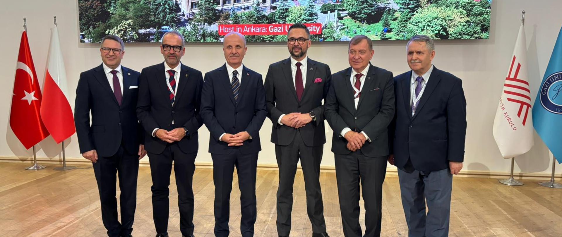 Pd ścianą na której wisi wielki ekran pokazujący stary budynek stoi sześciu mężczyzn w garniturach, trzeci od prawej minister Kulasek, za nimi pod ścianą po lewej flagi Polski i Turcji.