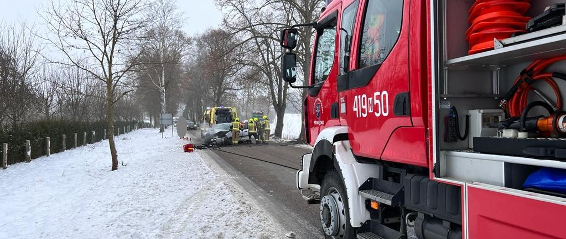 Zima. Na zdjęciu z prawej strony duży czerwony samochód strażacki stoi na drodze. W oddali na drodze rozbite auto i przy nim strażacy. Pobocza białe do śniegu.