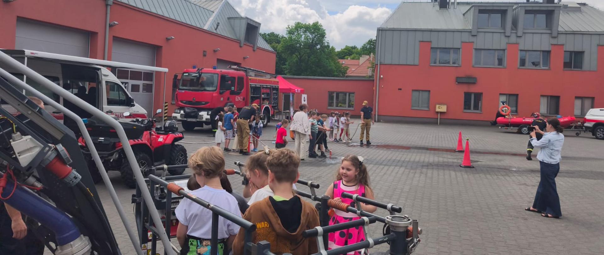 Grupa dzieci uczestniczy w pokazie strażackim na placu przed remizą; widoczne są wozy ratownicze, sprzęt strażacki oraz osoby obserwujące wydarzenie.