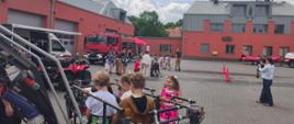 Grupa dzieci uczestniczy w pokazie strażackim na placu przed remizą; widoczne są wozy ratownicze, sprzęt strażacki oraz osoby obserwujące wydarzenie.