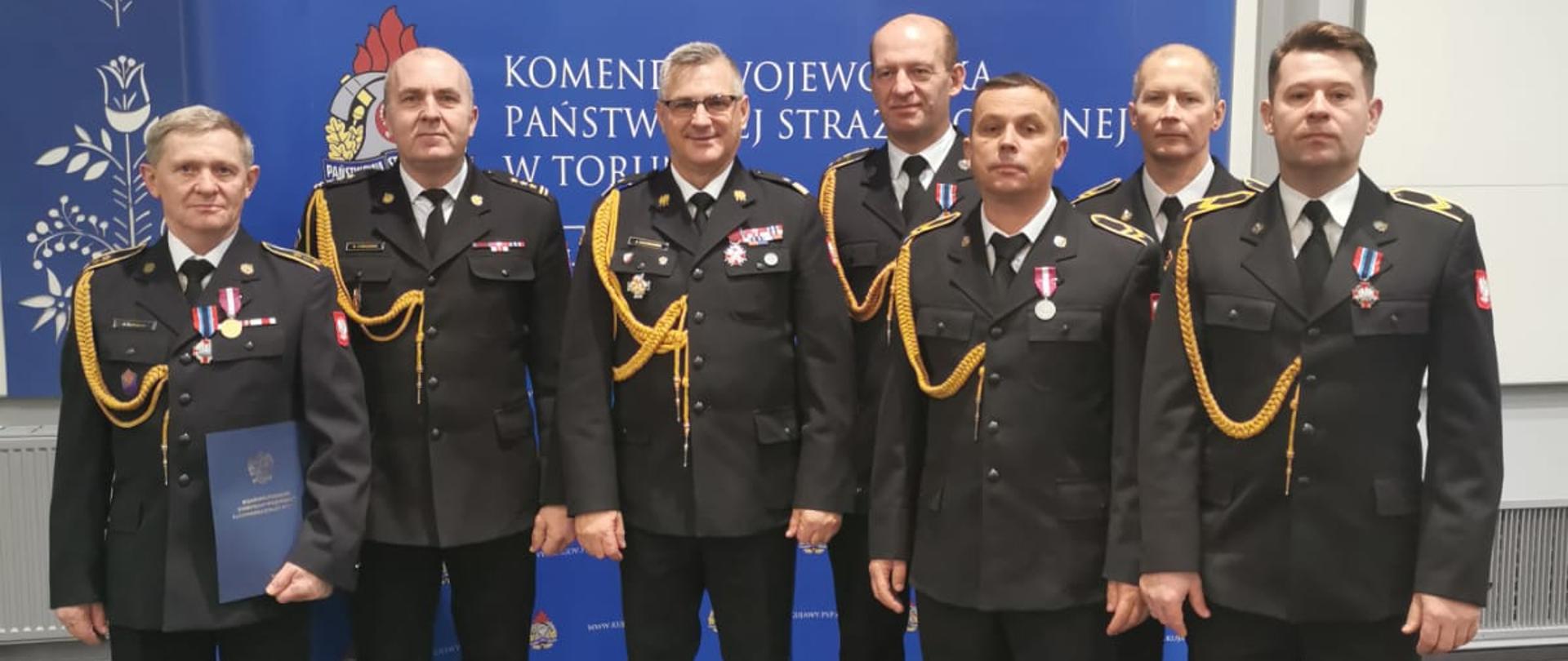Zdjęcie przedstawia Komendanta Wojewódzkiego PSP starszego brygadiera Jacka Kaczmarka ze strażakami PSP z Rypina, którzy dostali medale i wyróżnienia. Stoją na tle niebieskiego rolapu z logiem PSP. 
