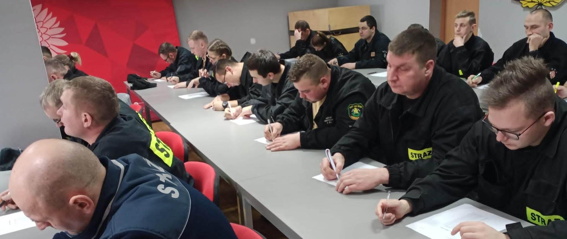 Recertyfikacja kpp. Świetlica w komendzie PSP w Rawiczu. Przy stołach siedzą druhowie OSP w umundurowaniu koszarowym, którzy uczestniczą w egzaminie recertyfikacyjnym. Zwróceni są w stronę okien. Strażacy rozwiązują test pisemny.