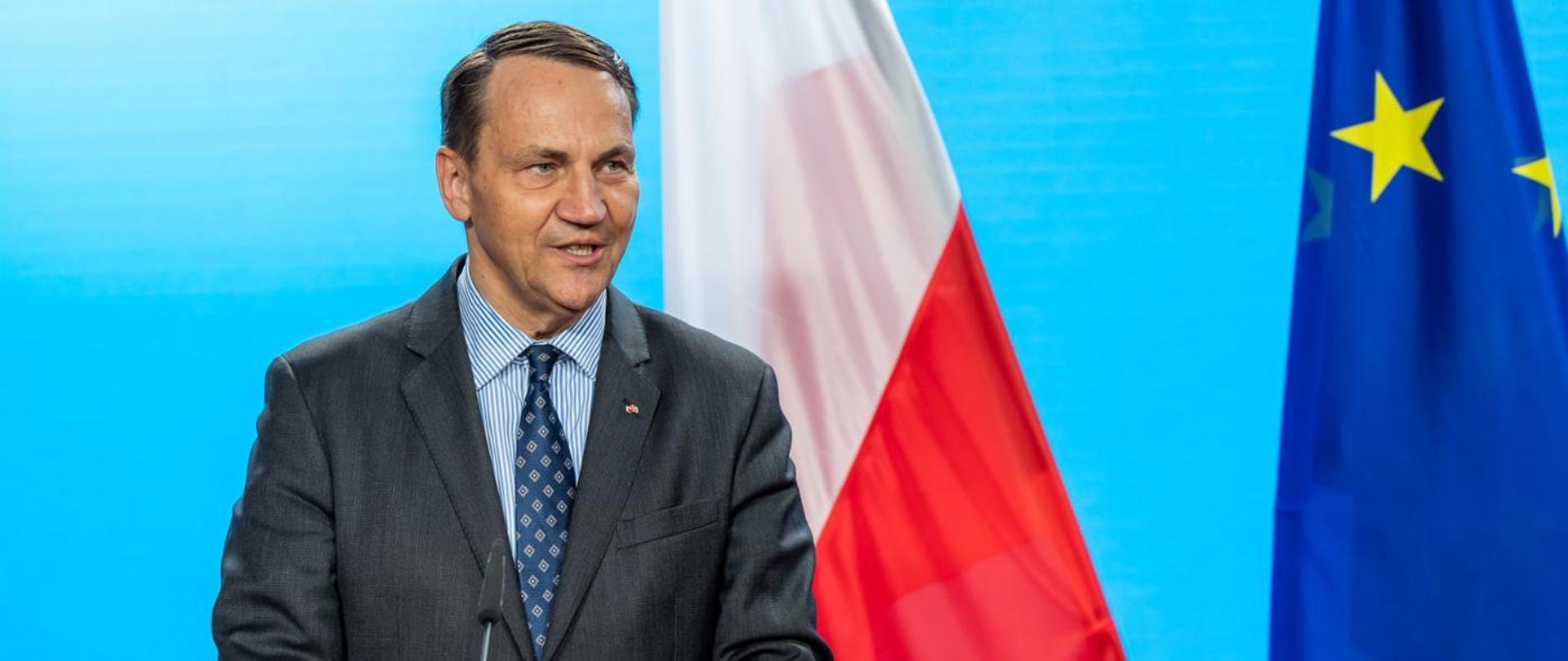 Mężczyzna w garniturze przemawia przed mikrofonem. Za nim stoją flagi Polska i Unii Europejskiej na błękitnym tle.