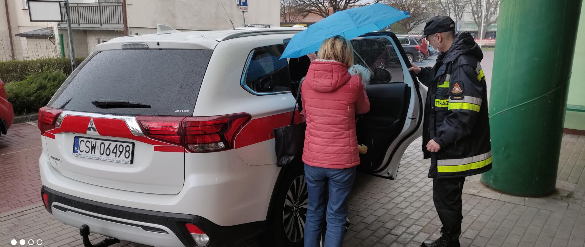 Na zdjęciu po prawej funkcjonariusz Komendy Powiatowej Państwowej Straży Pożarnej w Świeciu podczas pomocy osobie dowiezionej na szczepienie w wyjściu z samochodu. Tyłem na zdjęciu opiekun osoby dowiezionej na szczepienie.