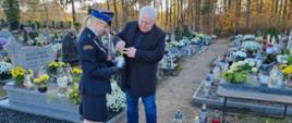 Zdjęcie przedstawia mężczyznę w cywilnym ubraniu i kobietę w mundurze strażackim, która kładzie znicz na grób. Stoją przed grobem, oddając hołd zmarłym.