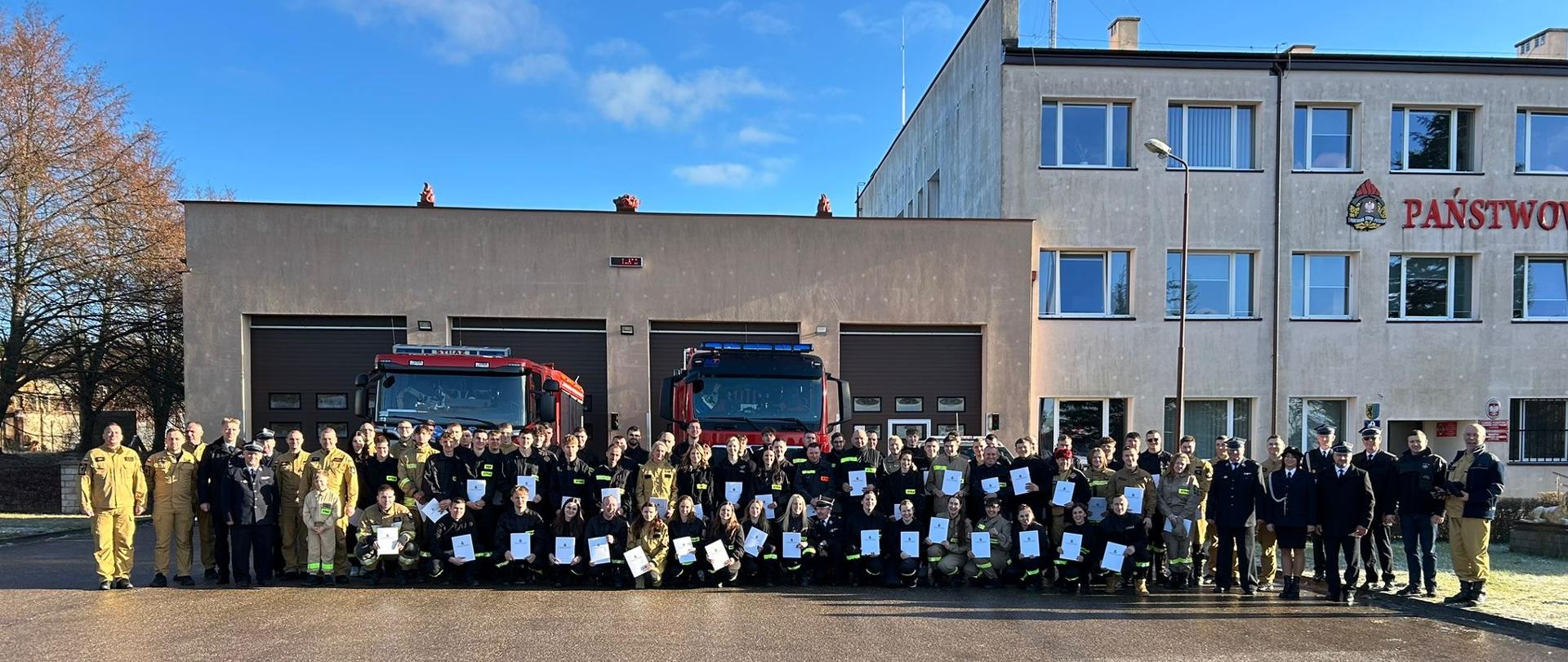 Uczestnicy szkolenia w szeregu. W tle budynek JRG oraz wozy strażackie.