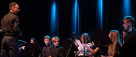 Zdjęcie przedstawia Zespół Muzyki Rozrywkowej PSM I stopnia w Jarocinie występujący na scenie podczas koncertu jubileuszowego. Na zdjęciu po lewej stronie znajduje się Pan Rafał Gubański dyrygujący zespołem uczniów.