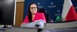 wiceminister Małgorzata Jarosińska-Jedynak uczestniczy w spotkaniu online przed komputerem, za nią flagi Polski i Unii Europejskiej
