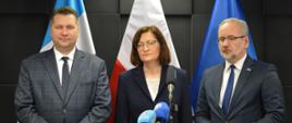 Minister Czarnek, kobieta ubrana na granatowo i minister Niedzielski stoją na tle ciemnoszarej ściany we wzór z kwadratów, za nimi flagi Polski i UE, przed nimi kilka mikrofonów na stojakach.