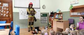 Na zdjęciu widzimy strażaka stojącego przed dziećmi i prezentującego maskę ochrony dróg oddechowych. Dzieci wsłuchane siedzą grzecznie. Strażak ubrany jest w odzież specjalną koloru piaskowego na głowie czerwony hełm z cyfrą 1. Zdjęcie ma miejsce w pomieszczeniu przedszkola widzimy szafki na których stoją książki.