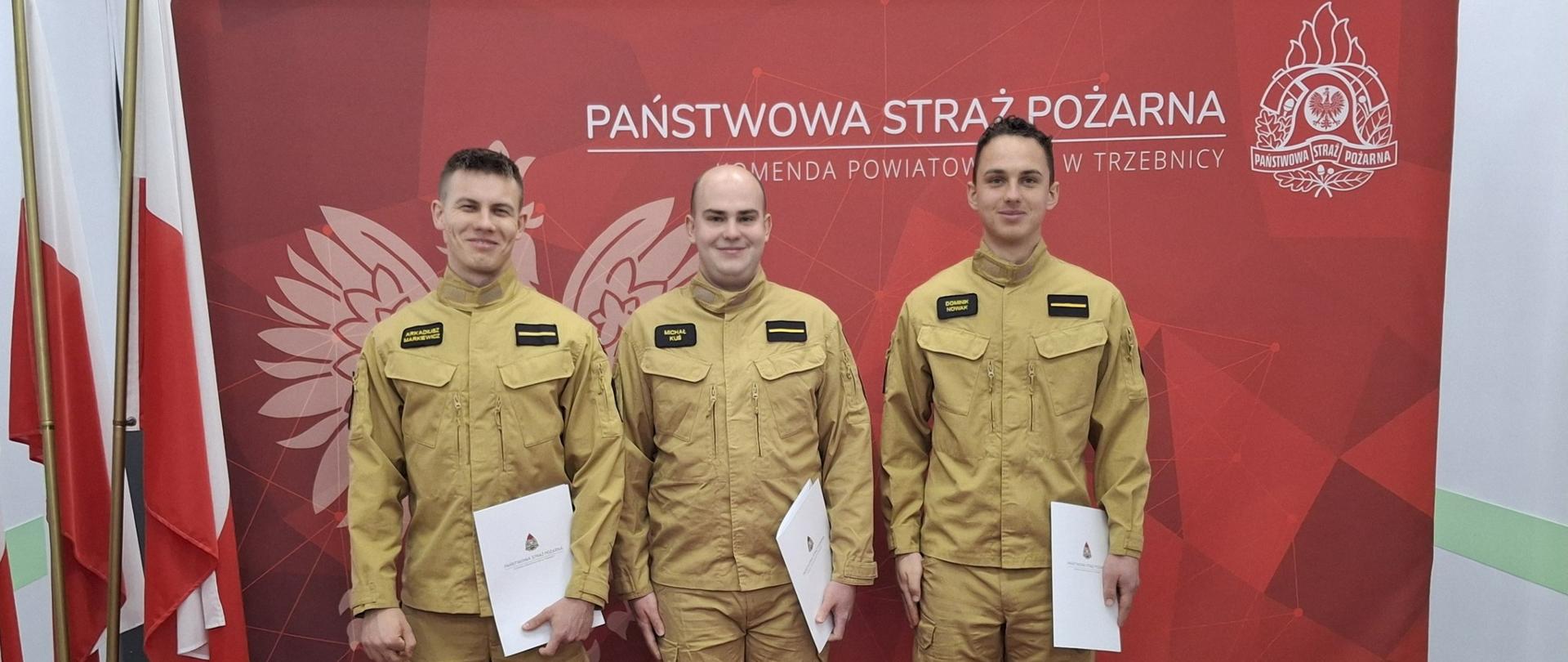 Awansowani strażacy na tle czerwonej ściany z logotypem Państwowej straży Pożarnej