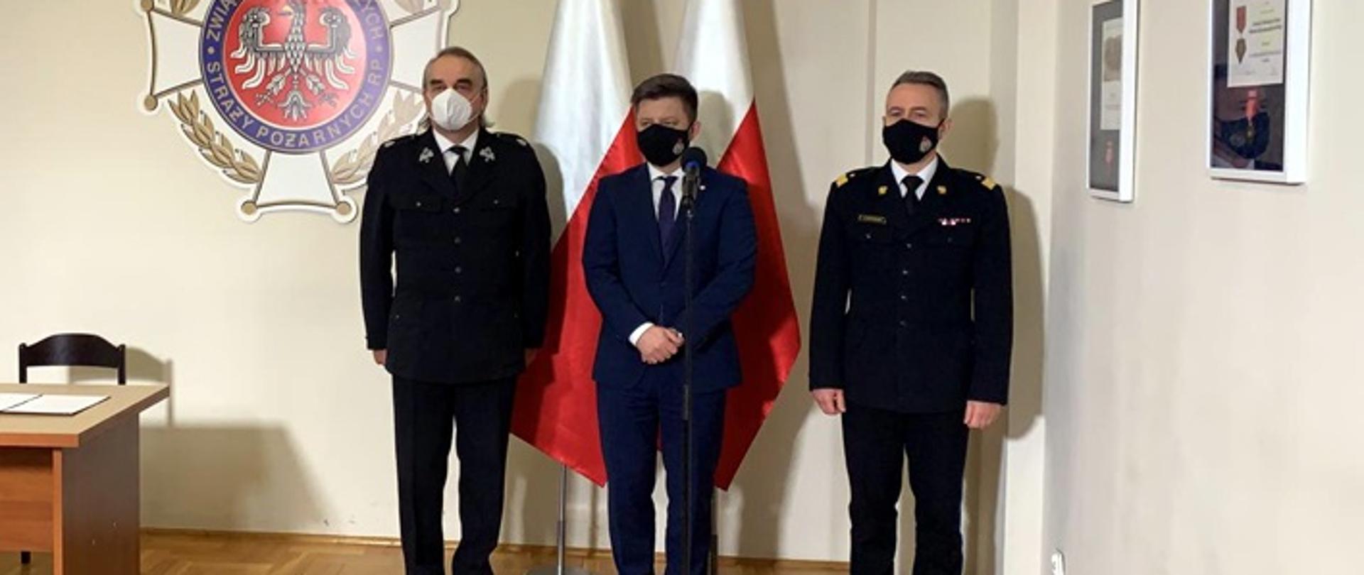 Na zdjęciu znajduje się 3 mężczyzn, dwóch ubranych w umundurowanie wyjściowe w kolorze czarnym, mężczyzna stojący pośrodku ubrany jest w granatowy garnitur. Za mężczyznami widać 2 flagi Polski oraz logo Związku OSP RP 