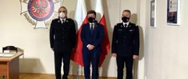 Na zdjęciu znajduje się 3 mężczyzn, dwóch ubranych w umundurowanie wyjściowe w kolorze czarnym, mężczyzna stojący pośrodku ubrany jest w granatowy garnitur. Za mężczyznami widać 2 flagi Polski oraz logo Związku OSP RP 