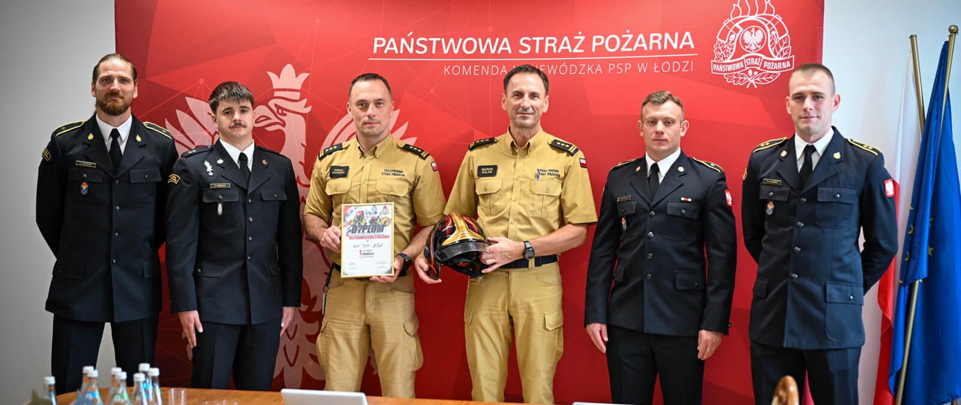 Pamiątkowe zdjęcie grupowe sześciu strażaków (czterech w mundurach wyjściowych i dwóch w piaskowych mundurach służbowych) na tle czerwonej ścianki Komendy Wojewódzkiej PSP w Łodzi. Na pierwszym planie widoczne są trzymane dyplomy i kask strażacki.