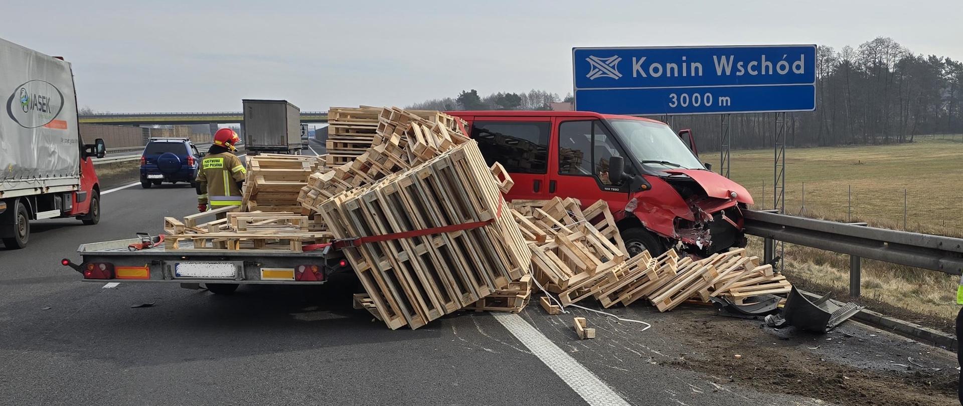 Samochód koloru czerwonego z lawetą przewożącą palety uderzył w barierę energochłonną. Palety częściowo leżą na drodze. Na drugim pasie jadą samochody. 