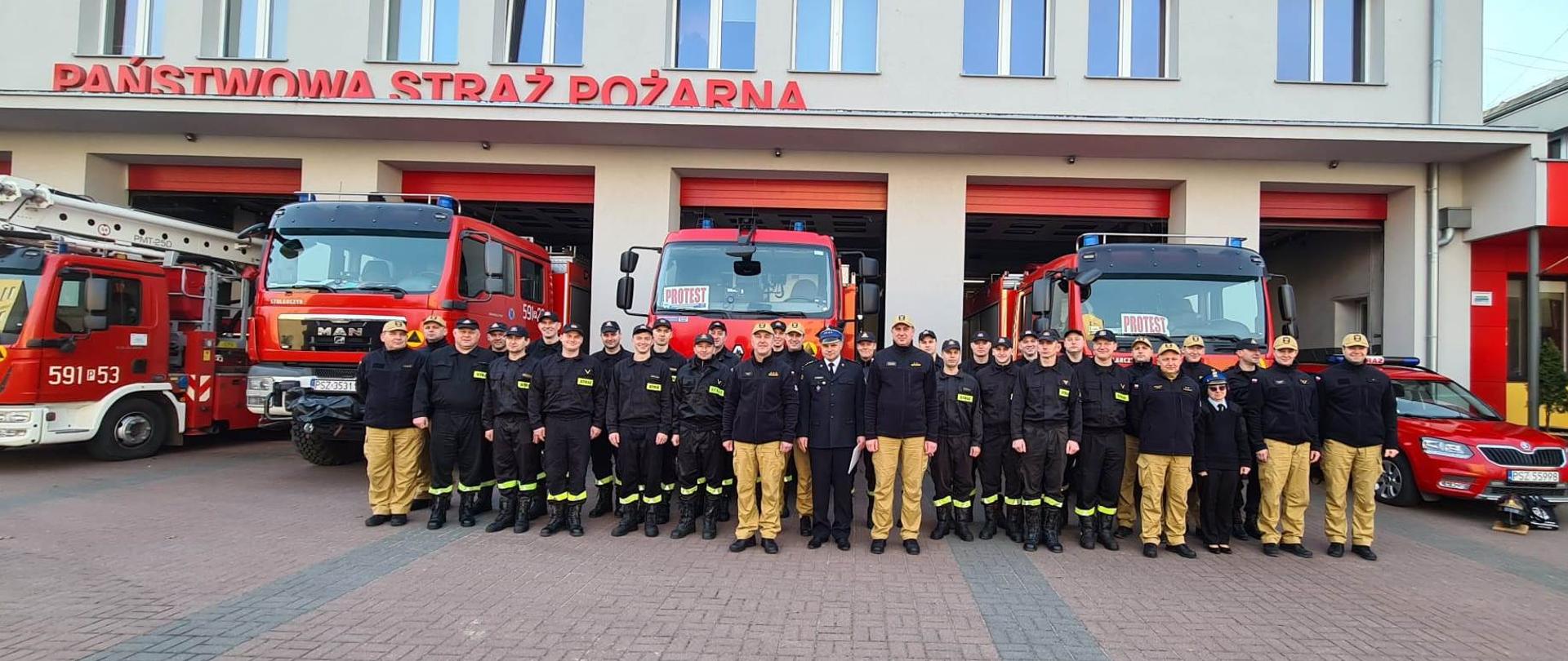 Widać strażaków na zdjęciu grupowym, za nimi samochody strażackie