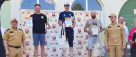 Fotografia przedstawia podium zwycięzców w kategorii mężczyzn wyścigu rowerowego MTB o puchar komendanta powiatowego. Po prawej i lewej stronie widać strażaków Państwowej Straży Pożarnej w ubraniu służbowym stojących obok podium. Wygrani prezentują dyplomy oraz statuetki.