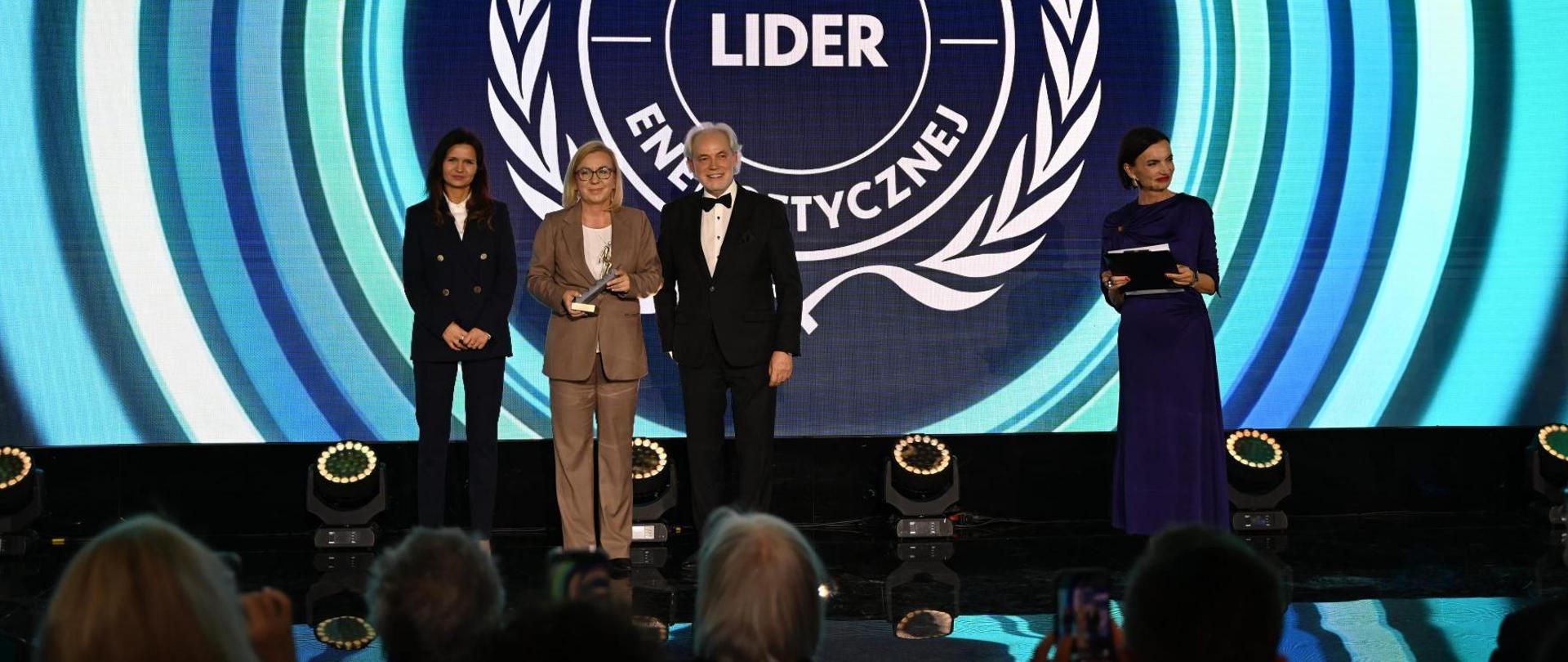 Wieczorna gala Polskiego Kongresu Klimatycznego. Na scenie stoją ministra klimatu i środowiska Paulina Hennig-Kloska w beżowy garniturze, obok niej po lewej stronie stoi kobieta, brunetka ubrana w ciemny garnitur, a po prawej siwy mężczyzna ubrany w ciemny garnitur. Ministra trzyma w ręku statuetkę - w kolorze złotym człowiek wspinający się po schodach. Na scenie za osobami baner z napisem Lider Transformacji Energetycznej. 