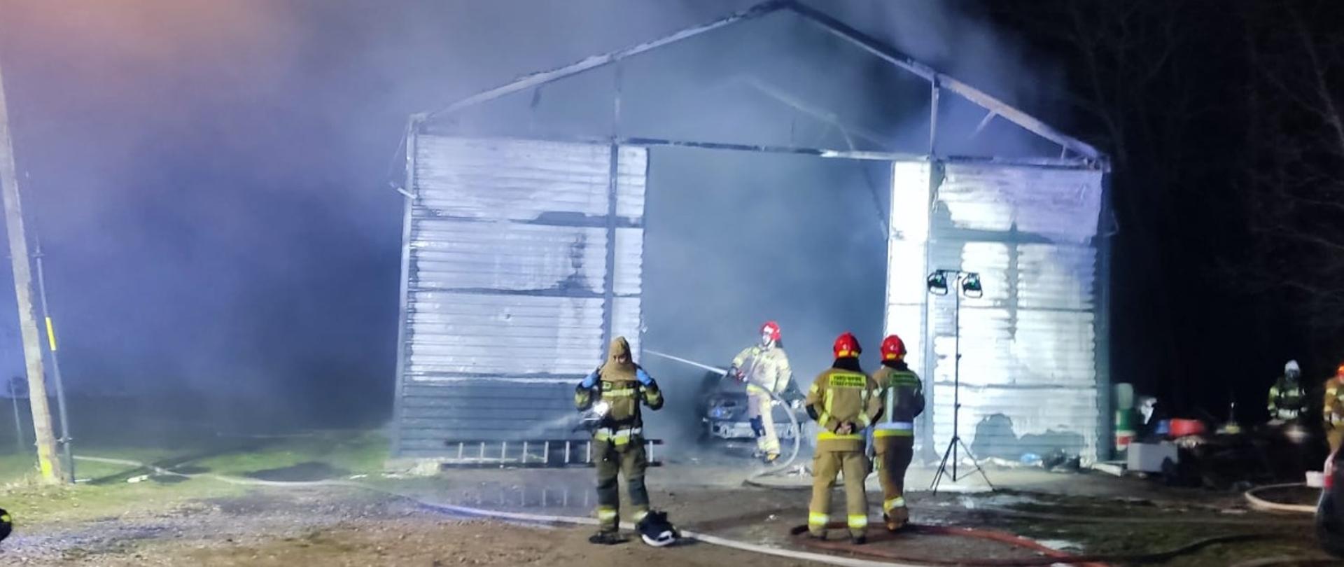 Zdjęcie wykonane w porze nocnej. Na zdjęciu widać spaloną halę warsztatową. W środku znajdują się spalone samochody. Przed halą stoją strażacy. Jeden z nich do środka leje wodę dogaszając palące się elementy wyposażenia. Strażacy ubrani są w ubrania specjalne w kolorze piaskowym. Na plecach założone mają butle z powietrzem. Przed warsztatem na ziemi znajdują się rozwinięte na ziemi linie gaśnicze zbudowane z węży pożarniczych. Po prawej stronie na zdjęciu widać ustawiony obok budynku samochód osobowy. Pojazd w kolorze ciemnym brązowym. 