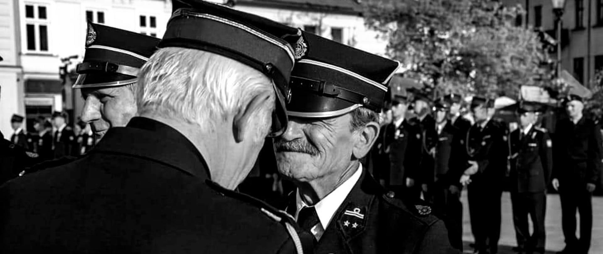 Widok z przodu. Druh Andrzej odznaczany odznaką św. Floriana przez kapelana małopolskich strażaków. W tle pododdział funkcjonariuszy Państwowej Straży Pożarnej. 