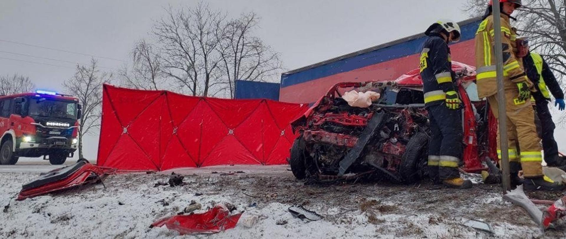 Wrak rozbitego pojazdu, koło niego strażacy i policja