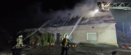 Zdjęcie wykonane w porze nocnej. Fotografia przedstawia strażaków PSP i OSP w ubraniu specjalnym koloru piaskowego ciemnego i czerwonych oraz białych hełmach na głowach, którzy przy pomocy prądów gaśniczych wody gaszą dach budynku mieszkalnego. Budynek znajduje się na środku kadru na drugim planie.