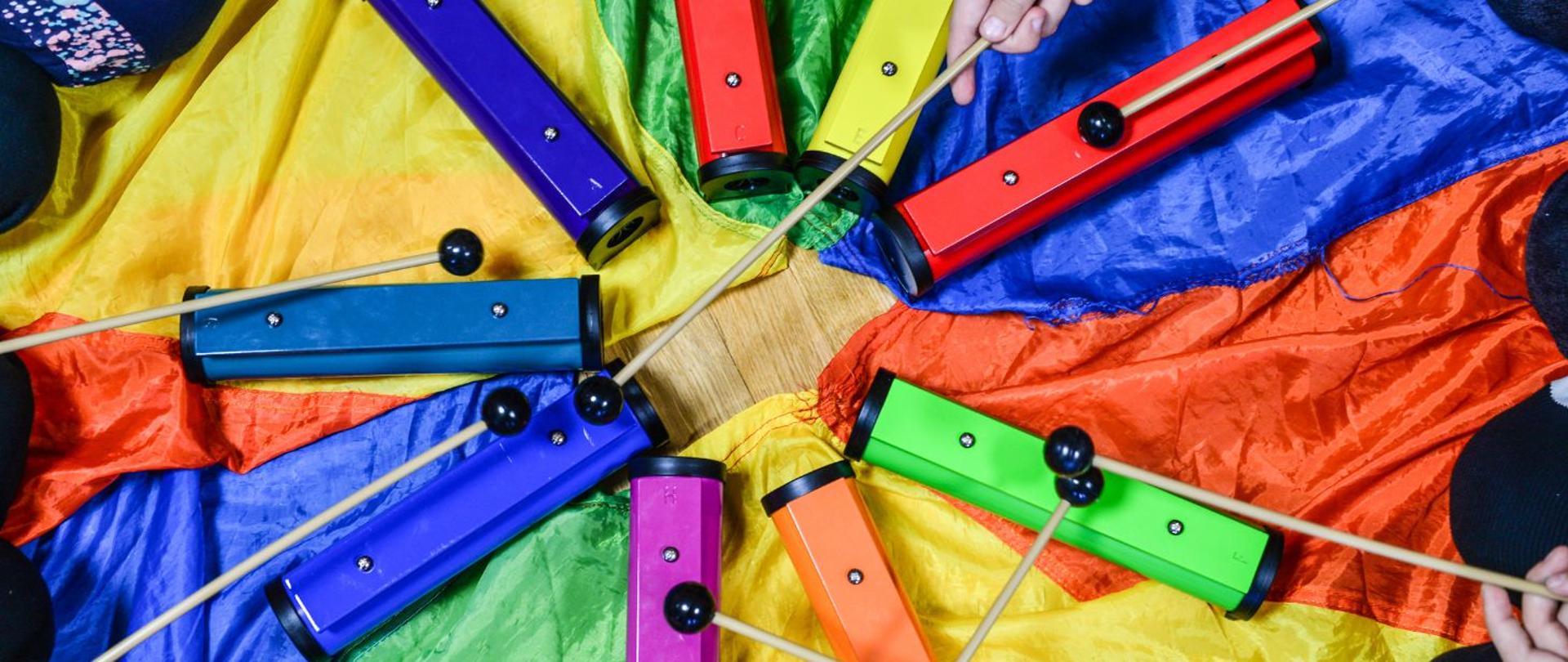 kolorowe zdjęcie przedstawia kolorowe sztabki metalofonu ułożone w kółko na tle chusty animacyjnej, oraz kolana i dłonie grających na sztabkach dzieci