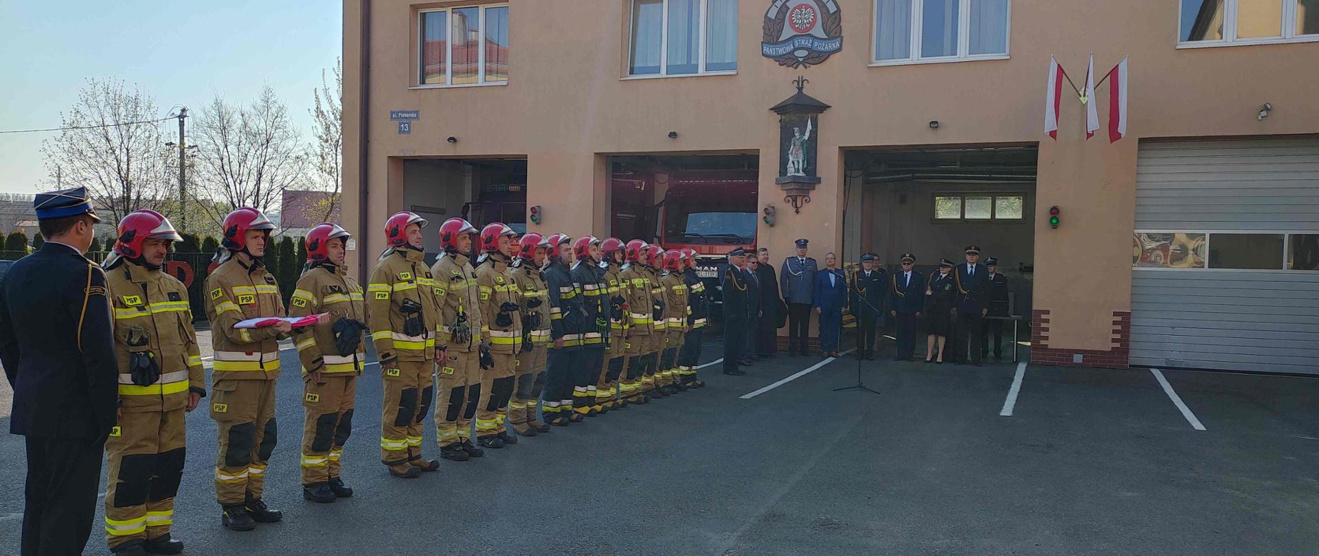  Zdjęcie przed uroczystością. Asfaltowy plac przed budynkiem jednostki ratowniczo – gaśniczej. Na budynku Logo Państwowej Straży Pożarnej, a pod nim figura św. Floriana. Budynek posiada duże bramy wjazdowe 3 otwarte i jedną zamkniętą. Nas środku ściany budynku znajdują się 3 flagi narodowe. Na placu stoją strażacy w szeregu pod kątem prostym do budynku. Pierwszy strażak (prowadzący uroczystość0 w mundurze wyjściowym następnie strażacy w ubraniach specjalnych. Pod budynkiem znajdują się Komendanci PSP w mundurach wyjściowych oraz zaproszeni goście. Jest jeden ksiądz, policjant, strażnicy miejscy oraz goście ubrani w garnitury. Na środku placu znajduję się mikrofon. Za zaproszonymi gośćmi w jednej z otwartych bram znajduję się stolik obsługi nagłośnienie wraz ze strażakiem obsługującym w mundurze wyjściowym. W dwóch pozostałych bramach znajdują się częściowo zasłonięte przez szereg strażaków samochody bojowe.