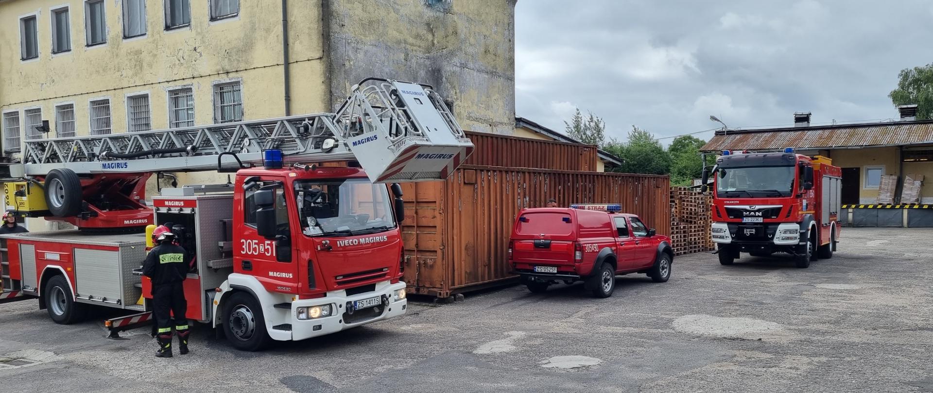 z lewej strony drabina mechaniczna rozkładana przy budynku i obsługujący ją strażacy, z prawej tyłem ustawiony samochód operacyjny Nissan pickup z obudowaną paką oraz samochód strażacki GBA marki MAN 