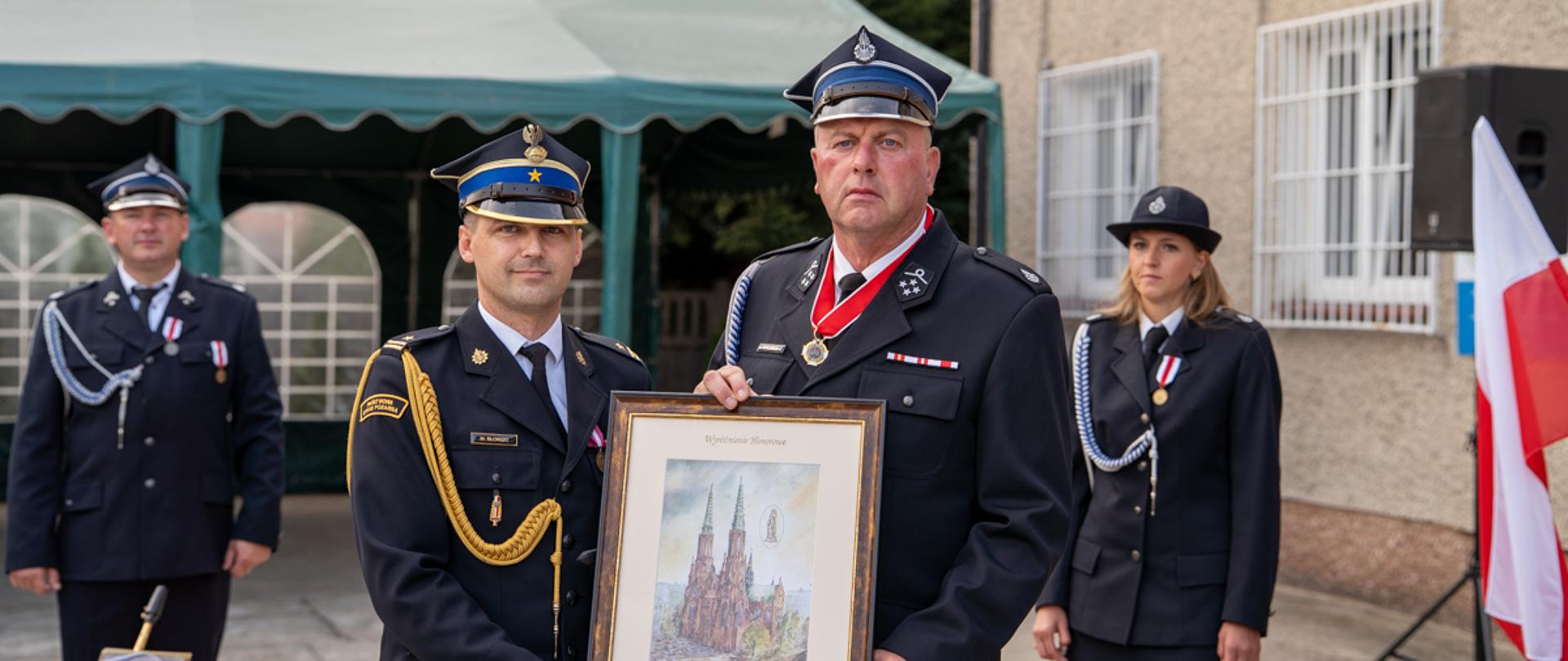 Dwóch mężczyzn w galowych mundurach strażackich pozuje do zdjęcia, trzymając dużą ramkę z obrazem przedstawiającym katedrę. Mężczyzna po lewej ma na sobie mundur z niebieskim pasem na czapce, złotym sznurem na ramieniu i rękawiczkami. Mężczyzna po prawej ma czerwony medal na szyi i poważny wyraz twarzy. W tle kobieta w mundurze oraz biało-czerwona flaga Polski.