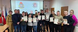 Na zdjęciu w dwóch rzędach stoją dzieci i młodzieży, laureaci konkursu oraz dorosłe osób, z czego czwórka z nich jest w mundurach. Dzieci i młodzież trzymają w dłoniach dyplomy oraz nagrody.