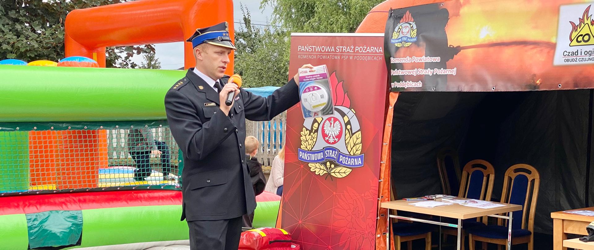 Strażak trzyma w ręce czujkę dymu i tlenku węgla pokazując ja uczestnikom. W drugiej ręce trzyma mikrofon. obok stoi pomarańczowy namiot, na którym jest plakat z logo PSP i żółte logo w kształcie ognia z napisem CO i napis Czad i Ogień obudź czujność. Przed namiotem sprzęt strażacki 