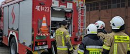 Zdjęcie przedstawia czterech strażaków stojących tyłem do obiektywu na betonowym placu przed otwartą skrytką sprzętową z tyłu czerwonego wozu strażackiego o numerze operacyjnym „401[D]23”. Trzech z nich ma na sobie piaskowo-brązowe mundury bojowe i białe kaski (jeden z napisem „OCHOTNICZA STRAŻ POŻARNA”, drugi „PAŃSTWOWA STRAŻ POŻARNA”), a jeden ubiór w kolorze ciemnogranatowym z żółtym napisem „STRAŻ”. W tle widoczny jest fragment starego, brązowego budynku garażowego pod pochmurnym niebem.