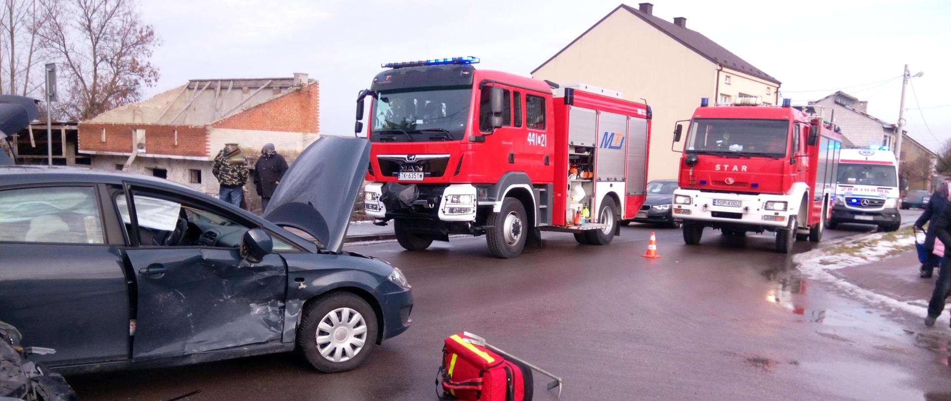 Zdjęcie przedstawia dwa samochody uczestniczące w wypadku. W tle widać uczestników zdarzenia dwa samochody pożarnicze oraz budynki mieszkalne.