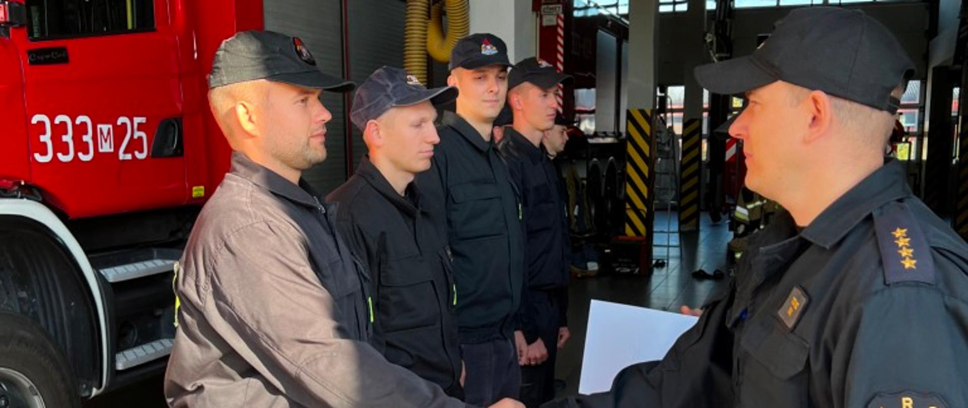 Zdjęcie wykonane na terenie obiektu jednostki ratowniczo – gaśniczej numer 3 w Radomiu. Na zdjęciu widać strażaków ustawionych w rzędzie. Przed nimi stoi zastępca dowódcy jednostki. Dokonuje oni wręczenia dyplomu strażakom działających w specjalistycznej grupy ratownictwa wysokościowego działającej w jednostce. Wszyscy strażacy ubrani są w ubrania koszarowe lub dowódczo – sztabowe w czarnym kolorze.
Za nimi znajduje się pojazd ratowniczo – gaśniczy w kolorze czerwonym z białymi i srebrnymi elementami zabudowy.
