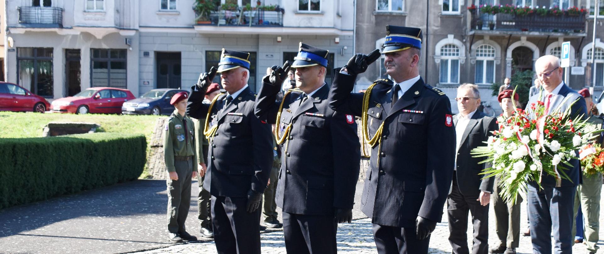 Strażacy w mundurach wyjściowych oddają hołd ofiarom II Wojny Światowej. Za nimi pozostali uczestnicy wydarzenia.