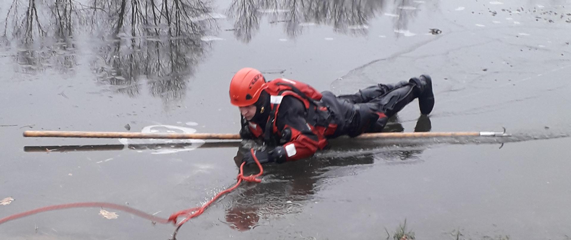 Zdjęcie przedstawia strażaka(pozoranta) w pełni zabezpieczonego leżącego na tafli lodu trzymającego w ręce linkę ratowniczą oraz leżącego na bosaku ratowniczym.