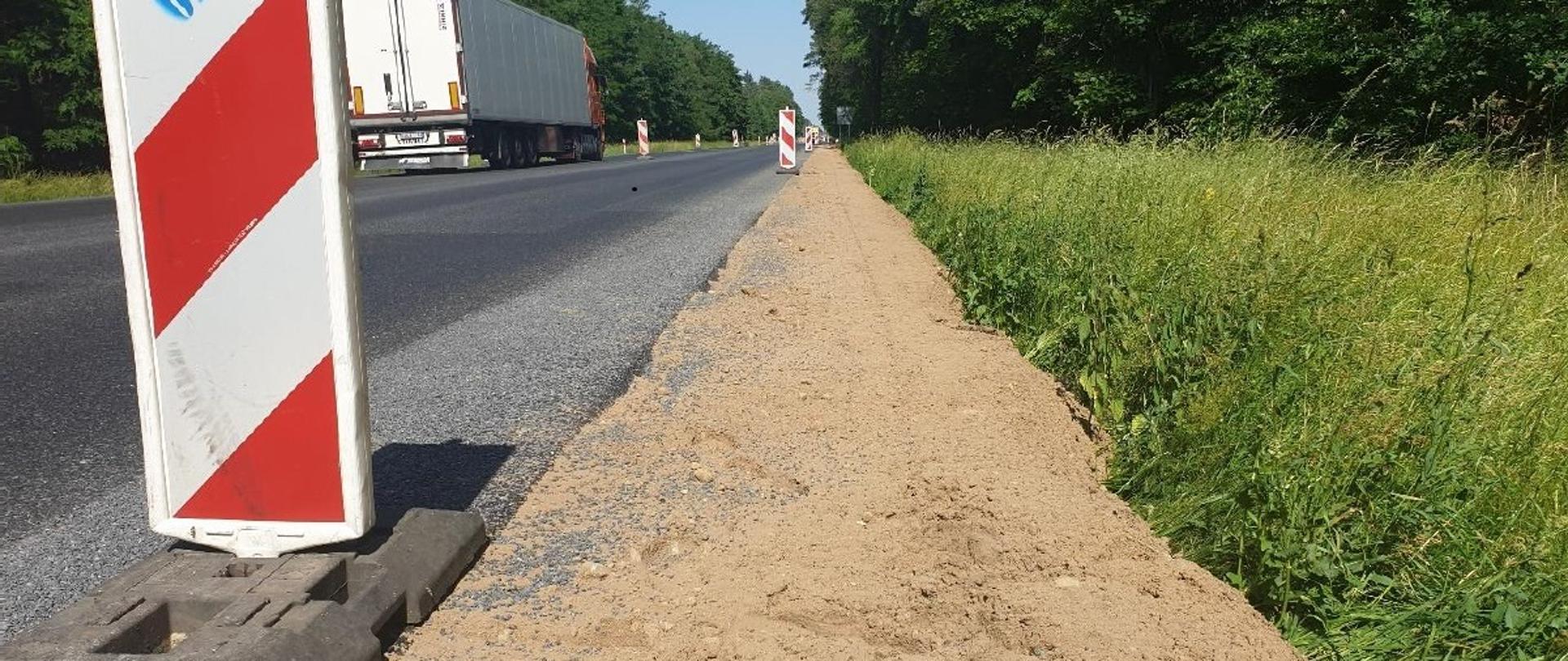 Zdjęcie z poziomu ziemi. Po lewej stronie droga, na której wzdłuż poboczy ustawione są znaki w biało-czerwone skośne pasy. Po prawej stronie zieleń.