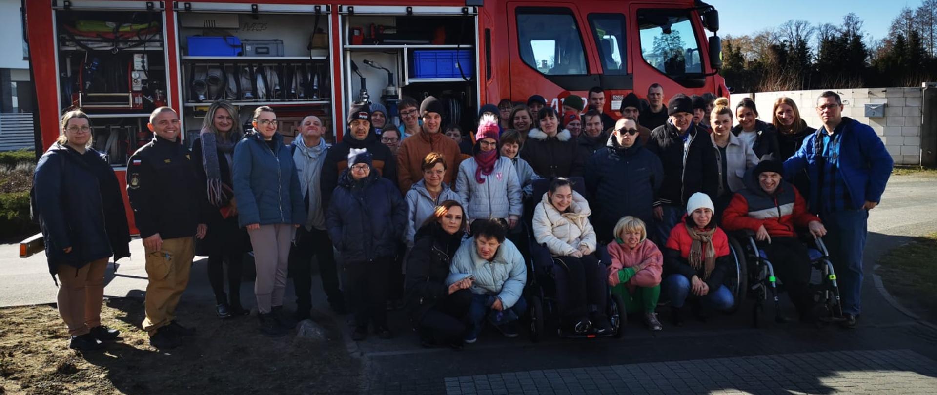 Zdjęcie zostało wykonane na zewnątrz. Na zdjęciu widzimy mężczyzn w mundurach straży pożarnej oraz podopiecznych warsztatów terapii zajęciowej na wspólnym zdjęciu.
W tle widoczny jest wóz strażacki.
