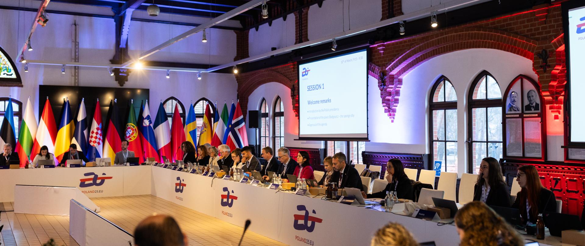 Sala z ludźmi, którzy siedzą przy prostokątnie ustawionych do siebie stołach. Za ich plecami widać flagi państw z całej Europy.