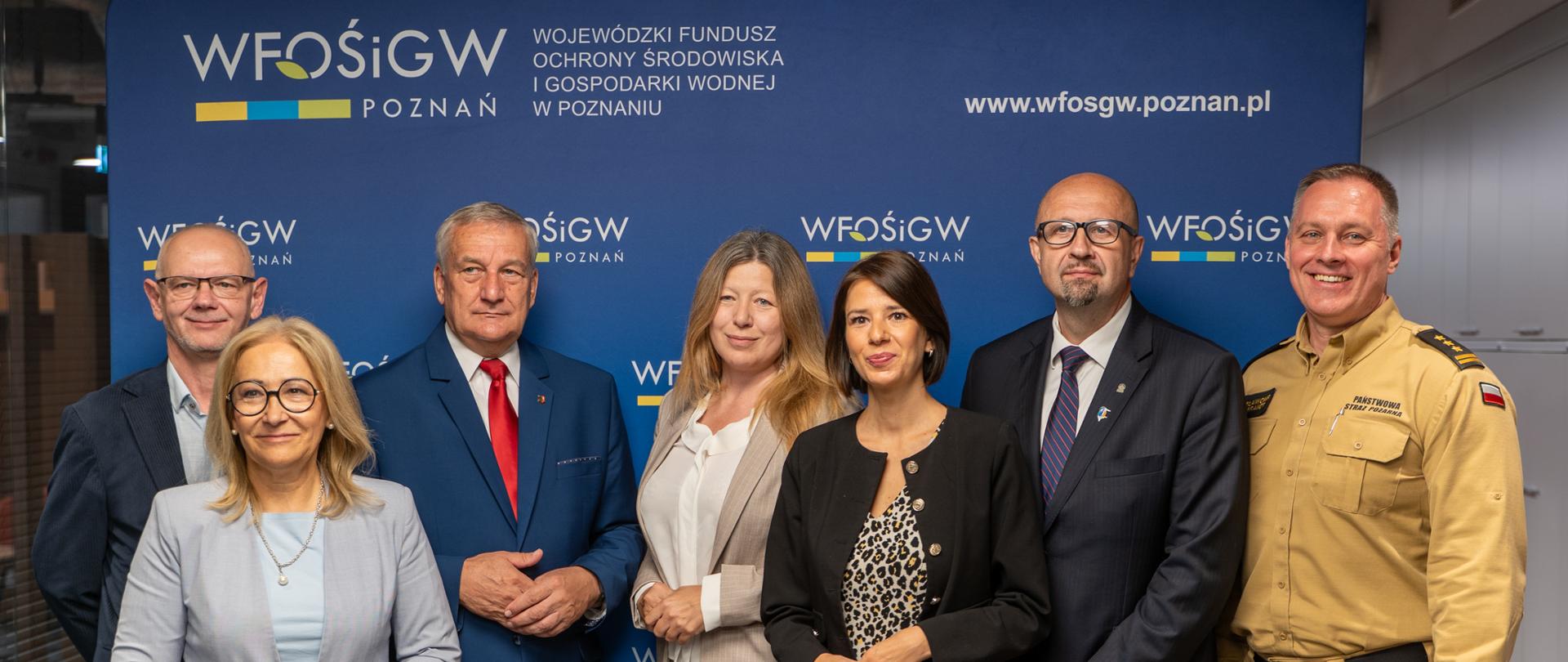 Grupa sześciu osób stoi do wspólnego zdjęcia na tle niebieskiej ścianki z logo i nazwą WFOŚiGW w Poznaniu oraz adresem strony internetowej funduszu. Uczestnicy są ubrani w stroje biznesowe, a jedna osoba ma mundur Państwowej Straży Pożarnej. Zdjęcie wykonano we wnętrzu biurowym podczas oficjalnego spotkania lub wydarzenia instytucjonalnego. Widoczne są napisy: „WFOŚiGW Poznań”, „Wojewódzki Fundusz Ochrony Środowiska i Gospodarki Wodnej w Poznaniu” oraz „www.wfosgw.poznan.pl”.