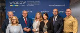 Grupa sześciu osób stoi do wspólnego zdjęcia na tle niebieskiej ścianki z logo i nazwą WFOŚiGW w Poznaniu oraz adresem strony internetowej funduszu. Uczestnicy są ubrani w stroje biznesowe, a jedna osoba ma mundur Państwowej Straży Pożarnej. Zdjęcie wykonano we wnętrzu biurowym podczas oficjalnego spotkania lub wydarzenia instytucjonalnego. Widoczne są napisy: „WFOŚiGW Poznań”, „Wojewódzki Fundusz Ochrony Środowiska i Gospodarki Wodnej w Poznaniu” oraz „www.wfosgw.poznan.pl”.