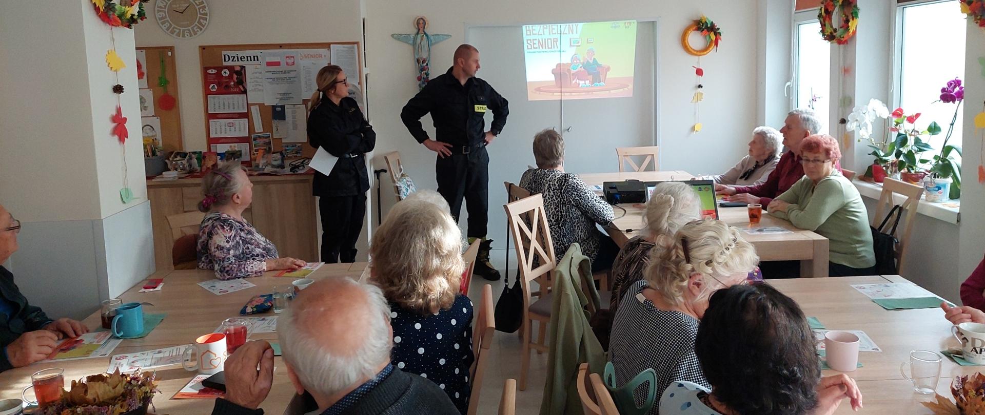 Umundurowana funkcjonariuszka i funkcjonariusz PSP stojący przed siedzącymi przy stolikach seniorami. W tle na ścianie grafika z prezentacji multimedialnej.